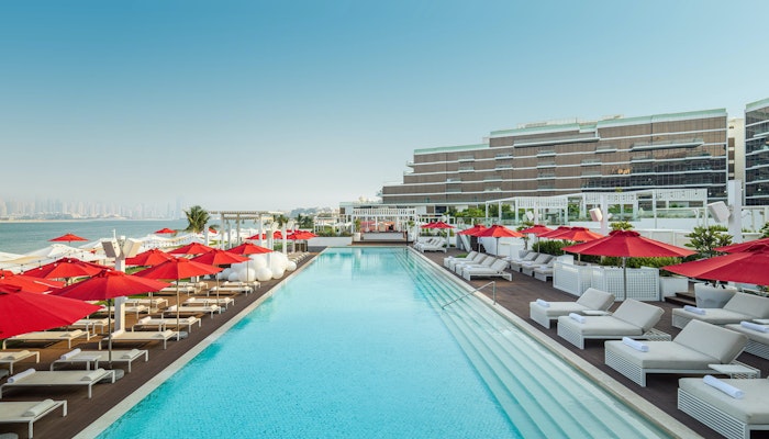 TH8 Palm Dubai Beach Resort - Vignette Collection Large pool with red umbrellas and sun loungers, overlooking the sea.