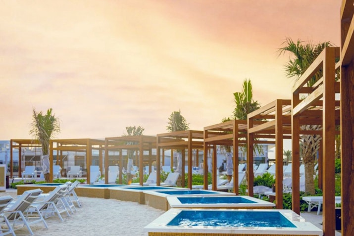 Wooden pergolas with lounges and small pools, set against a vibrant sunset sky.