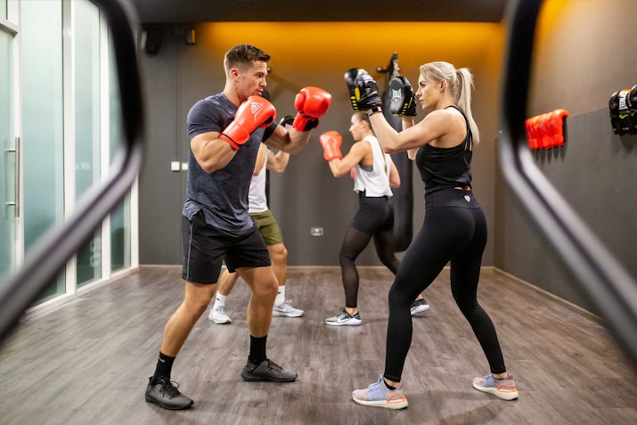 Two people in a gym engaged in a boxing training session, both wearing gloves and in a sparring stance. The woman is holding focus mitts while the man prepares to throw a punch. Another pair is training in the background, adding to the energy of the scene. The room has a modern design with boxing equipment mounted on the walls