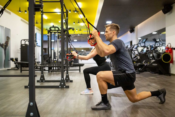 A man and a woman performing suspension training exercises in a gym, both kneeling and holding onto suspension straps while extending their arms forward. The gym features modern equipment and a brightly lit, spacious layout, with yellow and white ceiling accents.