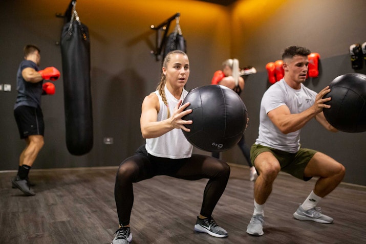 Instabody A man and woman performing squats while holding medicine balls in a fitness studio with a boxing area in the background. Other gym-goers are engaging in boxing exercises with punching bags in a well-lit space featuring a modern, motivational ambiance.