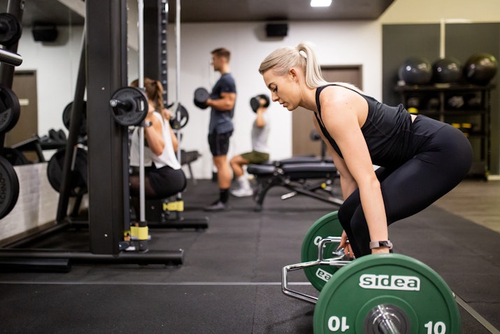 Instabody A woman in black workout attire performing a deadlift with green weight plates in a gym. She maintains proper form, with knees bent and back straight. In the background, other gym-goers engage in various exercises, including weightlifting and squats, adding to the dynamic atmosphere of the fitness space.
