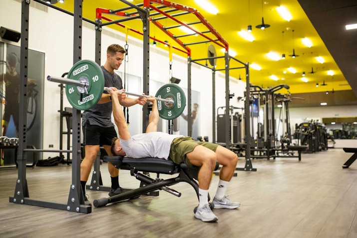 A man in athletic attire bench-pressing weights on a bench in a modern gym, assisted by a trainer standing behind the bar for support. The gym has various fitness equipment in the background, and the bright yellow ceiling adds a vibrant atmosphere