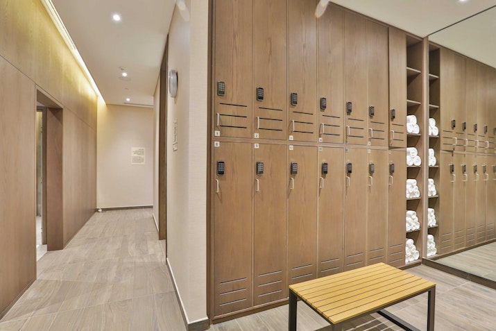 A modern locker room with light wood lockers, secure keypad locks, and a small bench in front. Towels are neatly stacked on open shelves beside the lockers, and soft lighting provides a clean, welcoming atmosphere