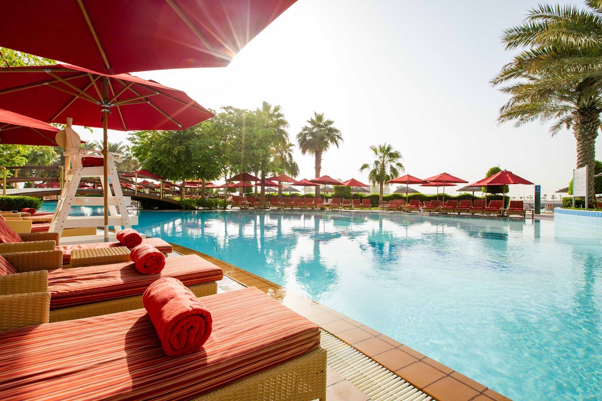 Poolside lounge chairs with red cushions and umbrellas, palm trees in the background.