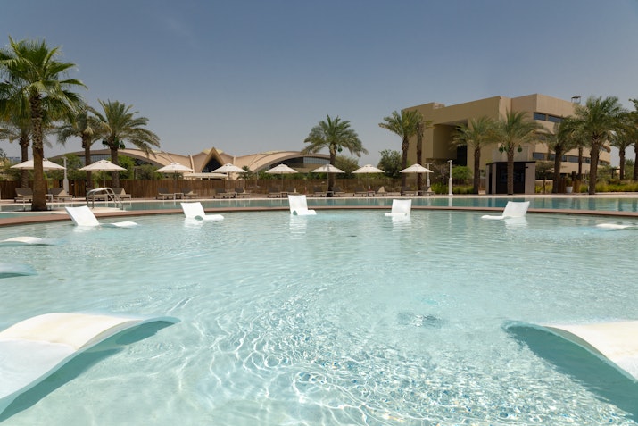 A serene pool with reclining lounge chairs partially submerged in the water, surrounded by palm trees and a resort building in the background.