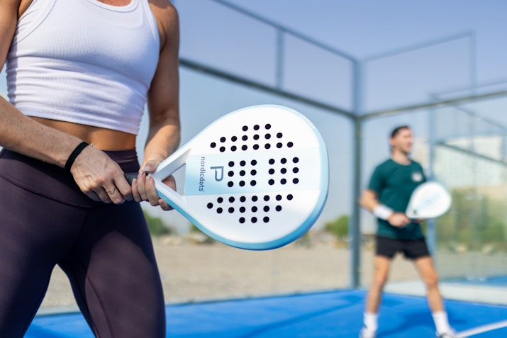 Close-up of a woman holding a padel racket while another person is in the background, on a padel court.