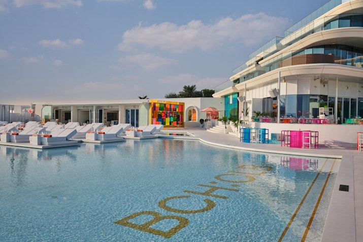 Pool area with white loungers, colorful decor, and modern building in the background.