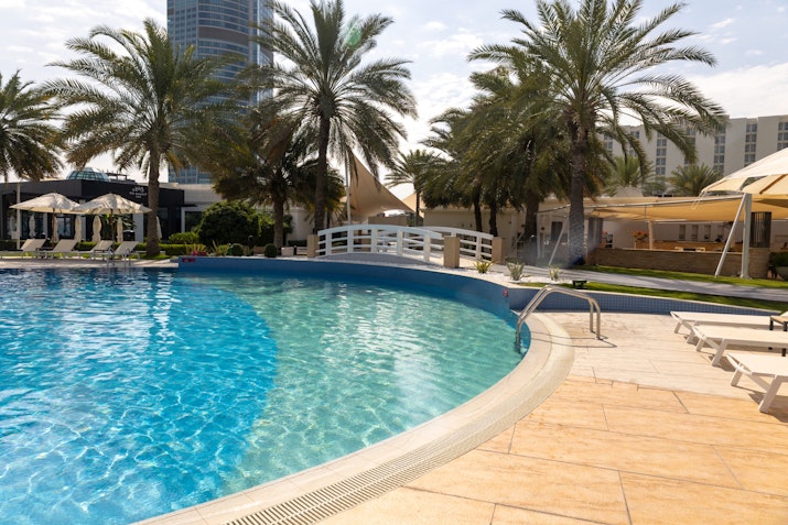 Curved pool with clear blue water, surrounded by palm trees, sun loungers, and a small bridge.