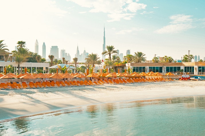 Beach with rows of orange loungers and umbrellas, with city skyline in the background.