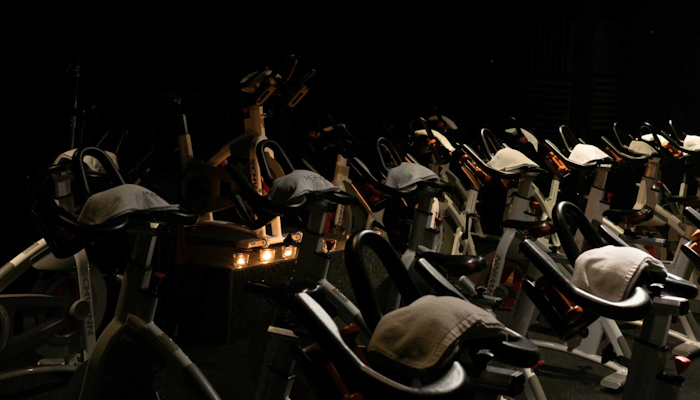 MOTION Cycling Rows of stationary bikes prepared for a spin class in a dimly lit room, with each bike having a white towel placed on its handlebars. The dark ambiance and low lighting create an intense, focused atmosphere, ideal for a high-energy indoor cycling workout