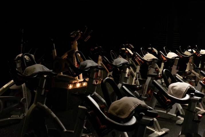 Rows of stationary bikes prepared for a spin class in a dimly lit room, with each bike having a white towel placed on its handlebars. The dark ambiance and low lighting create an intense, focused atmosphere, ideal for a high-energy indoor cycling workout