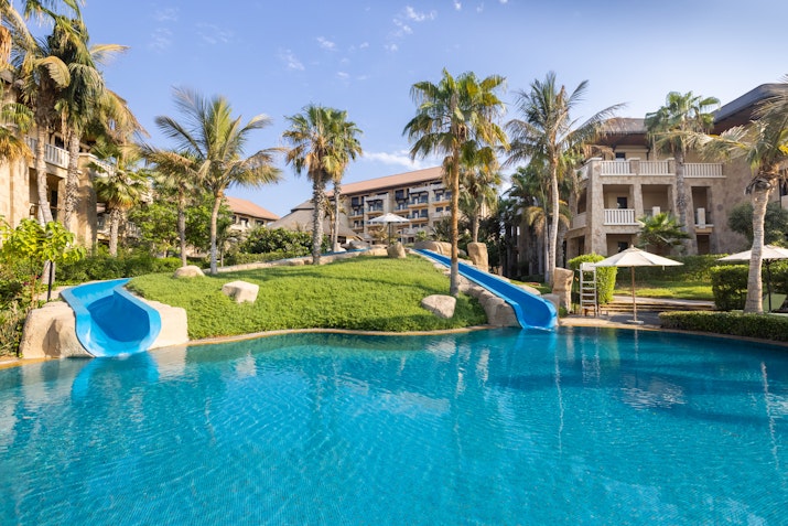 A pool with water slides, surrounded by lush greenery and palm trees, with resort buildings in the background.