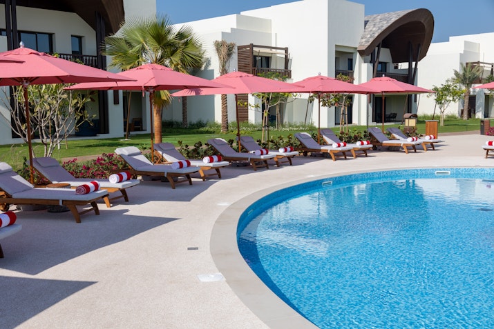 Anantara Mina Al Arab Ras Al Khaimah Resort Poolside lounge chairs under red umbrellas, with resort buildings in the background.