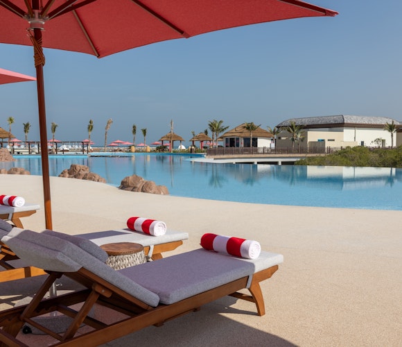Anantara Mina Al Arab Ras Al Khaimah Resort Lounge chairs with red umbrellas beside a serene pool area, overlooking a calm resort landscape.