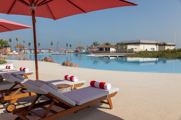 Lounge chairs with red umbrellas beside a serene pool area, overlooking a calm resort landscape.