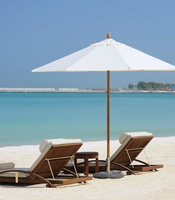 The St. Regis, Abu Dhabi Beach lounge chairs with a white umbrella, overlooking the turquoise sea and a clear sky.