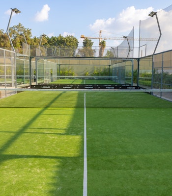 Mantra Padel A well-maintained outdoor padel court with vibrant green turf surrounded by glass walls and mesh fencing. The court is lit by overhead lights, and greenery and trees are visible beyond the fencing, adding a natural backdrop to the playing area. The sunny, clear sky complements the bright and inviting atmosphere of the court.