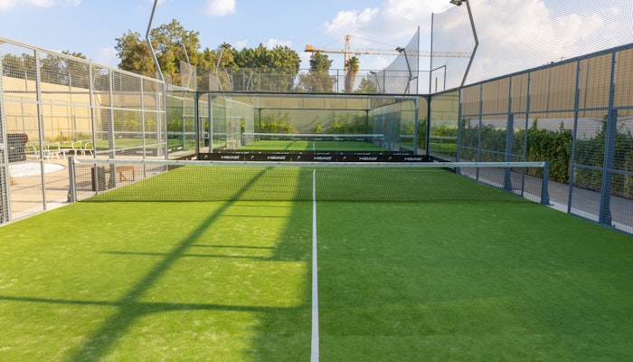 Mantra Padel A well-maintained outdoor padel court with vibrant green turf surrounded by glass walls and mesh fencing. The court is lit by overhead lights, and greenery and trees are visible beyond the fencing, adding a natural backdrop to the playing area. The sunny, clear sky complements the bright and inviting atmosphere of the court.