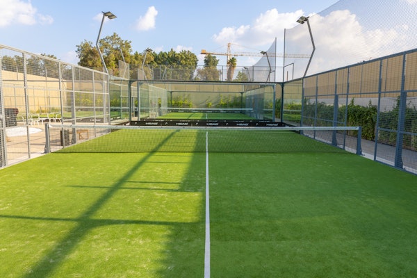 A well-maintained outdoor padel court with vibrant green turf surrounded by glass walls and mesh fencing. The court is lit by overhead lights, and greenery and trees are visible beyond the fencing, adding a natural backdrop to the playing area. The sunny, clear sky complements the bright and inviting atmosphere of the court.