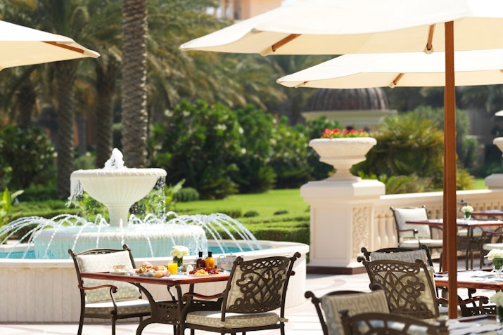Outdoor dining area with tables under umbrellas and a fountain in the background.