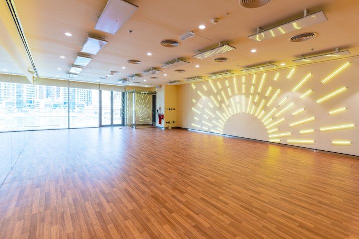 Empty fitness studio featuring wooden floors, large windows with city views, and a wall with sunburst LED lighting.