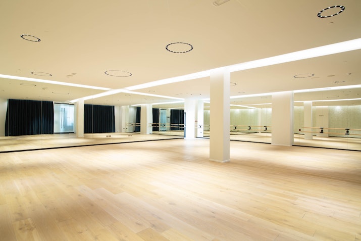 Spacious fitness studio with light wood floors, full-length mirrors, and a minimalist design featuring bright overhead lighting.