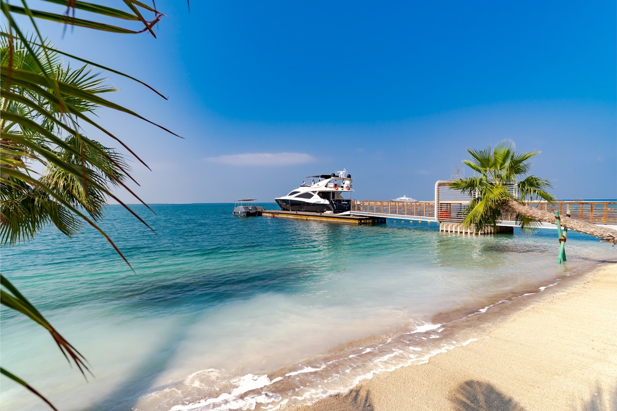 voco Monaco Dubai Scenic view of a sandy beach with clear blue waters, featuring a boat docked at a small pier, surrounded by palm trees.