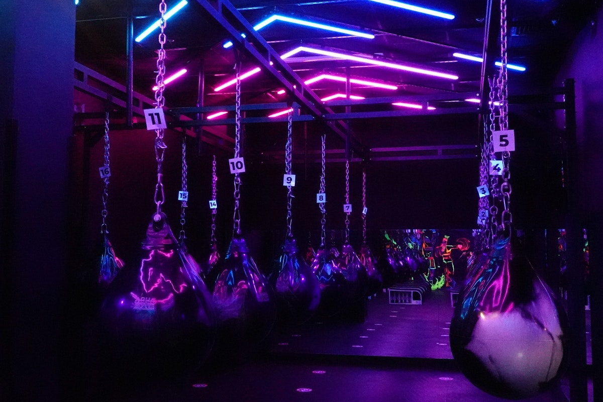 Boxica A boxing studio with large black and silver water punching bags hanging from chains, illuminated by vibrant neon pink, purple, and blue lights. The studio has a high-energy, futuristic atmosphere with reflective surfaces enhancing the colorful lighting