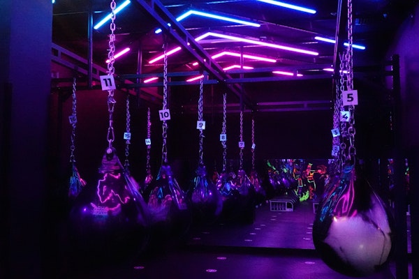 A boxing studio with large black and silver water punching bags hanging from chains, illuminated by vibrant neon pink, purple, and blue lights. The studio has a high-energy, futuristic atmosphere with reflective surfaces enhancing the colorful lighting
