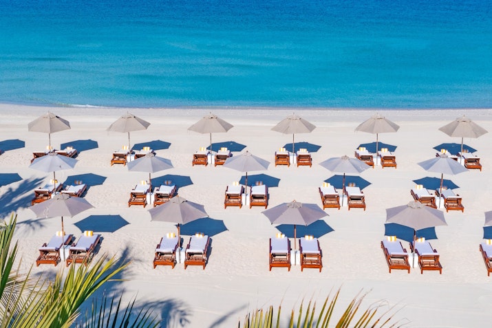 Aerial view of a sandy beach with sun loungers and umbrellas arranged neatly along the shoreline, next to calm blue waters.