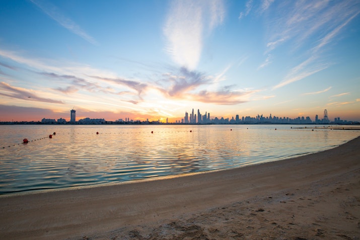 Sunset over the skyline with calm water and beach in the foreground.