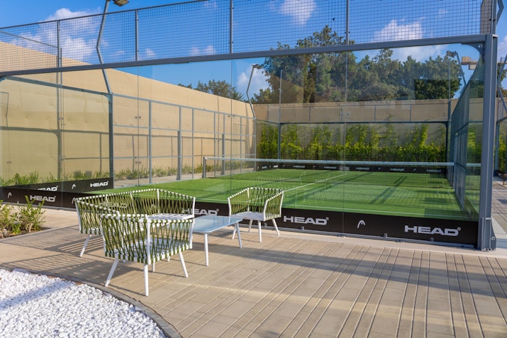 Mantra Padel A well-maintained outdoor padel court surrounded by glass walls, featuring green turf and ample lighting for evening play. Adjacent to the court is a stylish seating area with modern white woven chairs and a small table, placed on a light wooden deck with a decorative white stone border. The setup provides a comfortable spot for players to relax and spectate in a peaceful outdoor setting