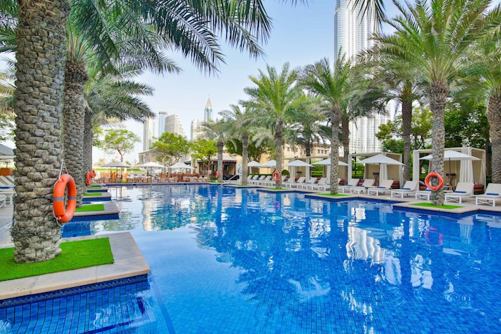 The Palace Downtown A luxury pool with palm trees and lounge chairs, set against a city backdrop.