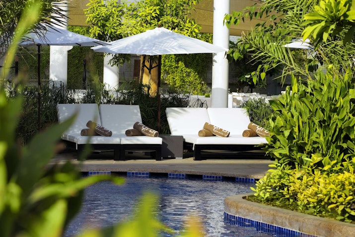 Conrad Dubai  Lounge chairs by a pool, surrounded by lush greenery and shaded by umbrellas in a tranquil setting.