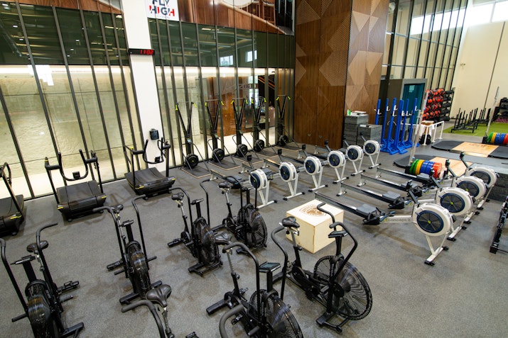 Well-equipped gym featuring multiple rowing machines, air bikes, and other cardio equipment, set against a modern interior with large glass walls.