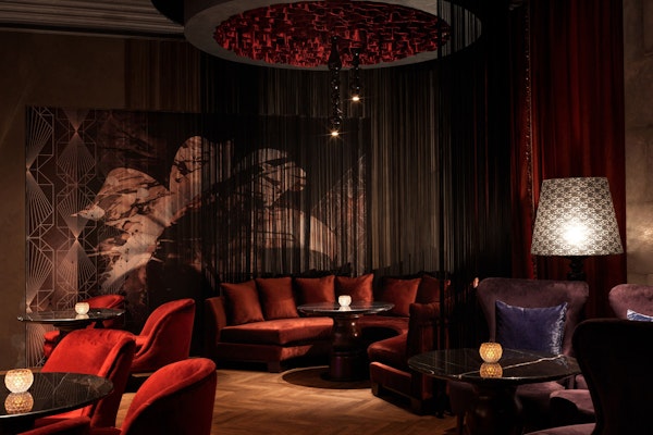 Luxurious, dimly-lit dining lounge with plush red and velvet seating, ornate chandeliers, and atmospheric decor, creating an intimate and sophisticated setting for dinner and a show.