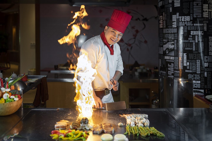 A smiling chef in a red hat performing a teppanyaki show with flames rising from the grill, surrounded by colorful vegetables and fresh ingredients, creating a lively and entertaining dining atmosphere.