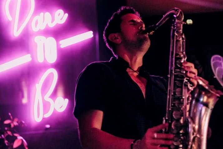 A saxophonist passionately playing in a dimly lit venue, with a neon 'Dare to Be' sign glowing pink in the background, setting a vibrant and lively atmosphere