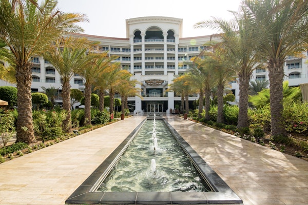 Waldorf Astoria A pathway with palm trees leading towards a building, with a fountain flowing through the center.