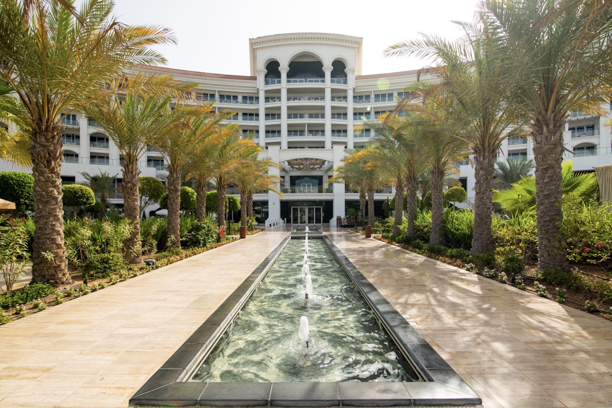 Waldorf Astoria A pathway with palm trees leading towards a building, with a fountain flowing through the center.