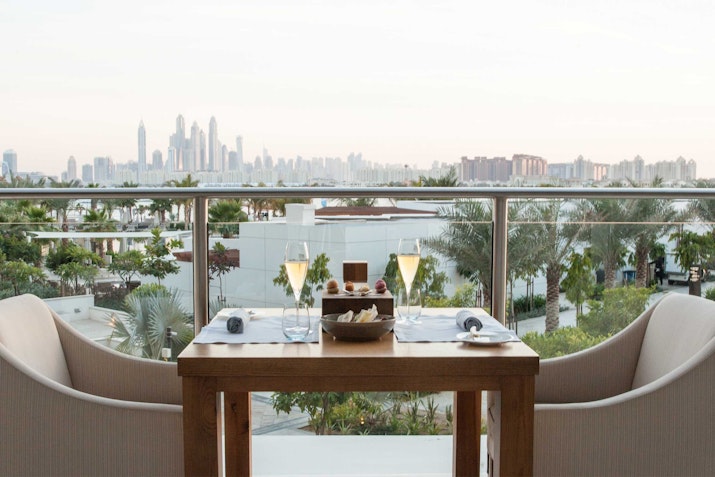 A dining table set for two with glasses and cutlery, overlooking a city skyline from a balcony.