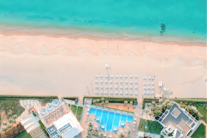 An aerial view of a beautiful beach with a turquoise sea, showcasing sun loungers and a swimming pool near a beachfront hotel.