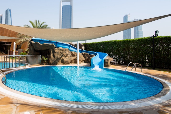 A pool with a water slide, surrounded by modern buildings and a shade sail.