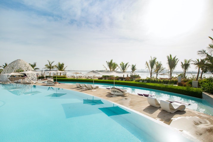 A scenic view of a poolside with lounge chairs, palm trees, and a beach in the background.