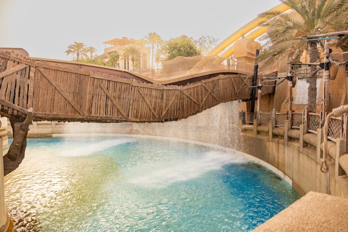 A water park scene featuring a wooden bridge over a turquoise pool with cascading waterfalls, surrounded by tropical landscaping and water slides in the background