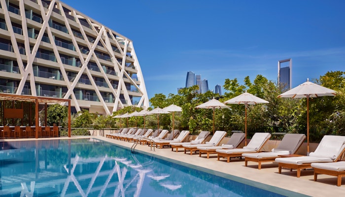 The Abu Dhabi EDITION Poolside with loungers and umbrellas beside a modern hotel building against a clear blue sky.
