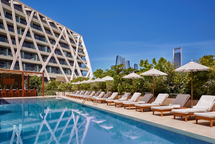 Poolside with loungers and umbrellas beside a modern hotel building against a clear blue sky.