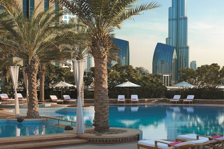 Shangri-La Dubai A luxurious swimming pool surrounded by palm trees and lounge chairs, with modern buildings in the background.