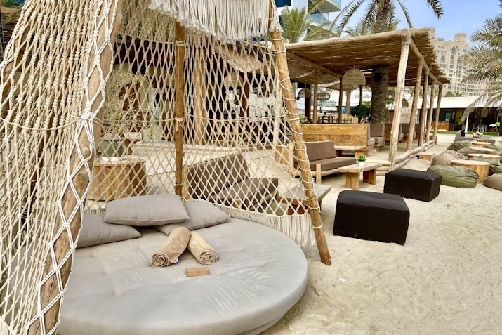 Cozy beach seating area featuring a large round cushioned lounge surrounded by wooden furniture and tropical decorations.